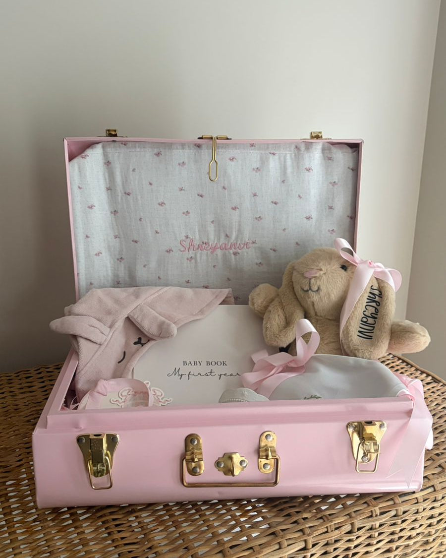Pink keepsake trunk with baby items and a teddy bear on a wicker surface.